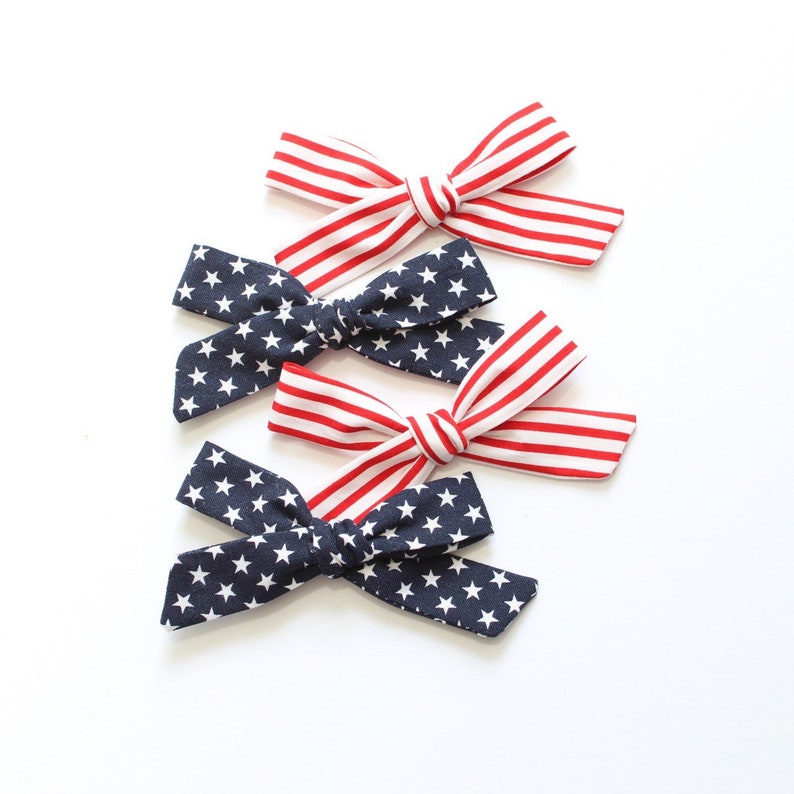 May include: Three fabric hair bows in red, white, and blue with a star pattern. The bows are arranged in a stack, with the red, white, and blue bow on top, followed by a blue star bow, and then another red, white, and blue bow.