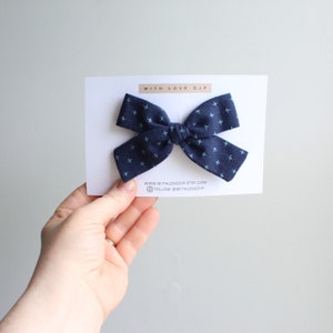 Puede incluir: Un lazo para el pelo azul marino con un patr&oacute;n de cruces blancas. El lazo est&aacute; en una tarjeta blanca con el texto "WITH LOVE DJP" e informaci&oacute;n del sitio web.