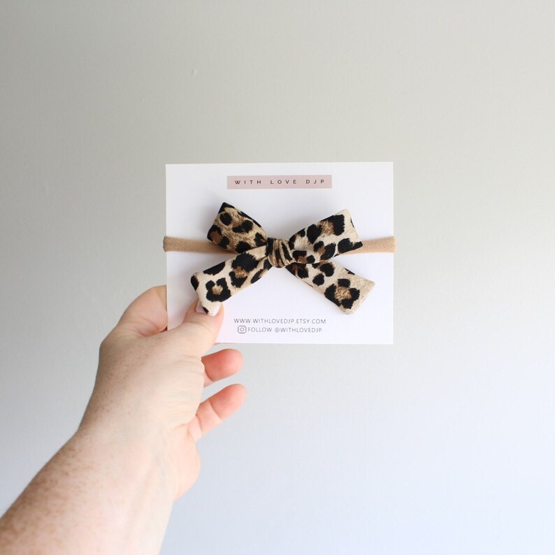 Puede incluir: Un lazo de tela con estampado de leopardo marr&oacute;n y negro en una diadema el&aacute;stica de color nude. La diadema se muestra en una tarjeta blanca con el texto "WITH LOVE DJP" y "www.withlovedjp.etsy.com" y "FOLLOW @withlovedjp".