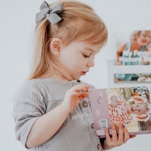 Puede incluir: Una ni&ntilde;a peque&ntilde;a con cabello rubio que lleva una camisa gris con botones y un lazo gris en el pelo est&aacute; leyendo un libro con una portada rosa. El libro tiene una foto de un ni&ntilde;o sonriente en la portada y la palabra "Mam&aacute;" impresa en la portada.