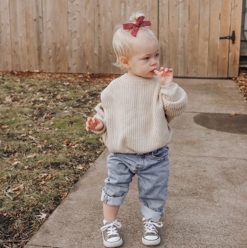 Puede incluir: Un ni&ntilde;o peque&ntilde;o con un su&eacute;ter de punto beige claro, jeans azul denim y zapatillas blancas. El ni&ntilde;o tiene un lazo burdeos en el pelo y est&aacute; de pie en una acera.