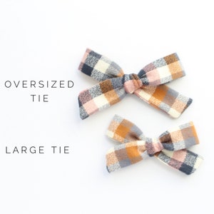 May include: Two plaid hair bows, one larger than the other. The larger bow is labeled "Oversized Tie" and the smaller bow is labeled "Large Tie". Both bows are made of a soft, plaid fabric in shades of pink, brown, white, and gray.