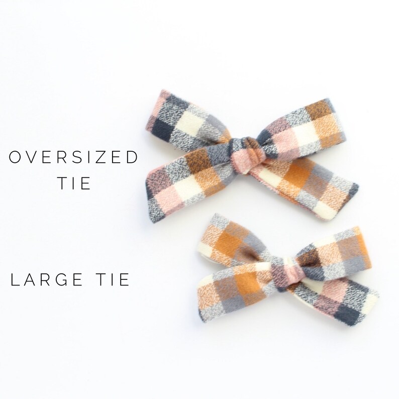 May include: Two plaid hair bows, one larger than the other. The larger bow is labeled "Oversized Tie" and the smaller bow is labeled "Large Tie". Both bows are made of a soft, plaid fabric in shades of pink, brown, white, and gray.