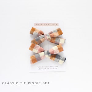 May include: Set of two plaid hair bows in a classic tie piggie style. The bows are made of a soft, textured fabric in shades of orange, pink, white, and gray. The bows are tied with a gray ribbon and are perfect for adding a touch of whimsy to any outfit.