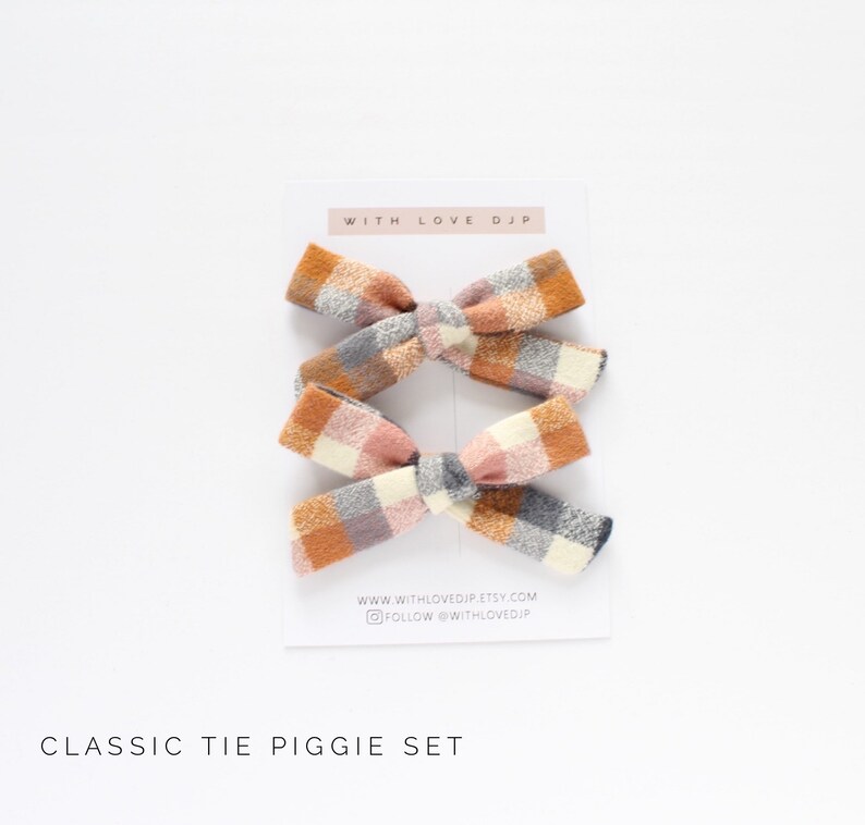 May include: Set of two plaid hair bows in a classic tie piggie style. The bows are made of a soft, textured fabric in shades of orange, pink, white, and gray. The bows are tied with a gray ribbon and are perfect for adding a touch of whimsy to any outfit.