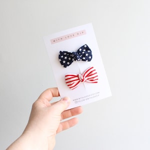 May include: Two small bow ties, one navy blue with white stars and the other red and white striped, are displayed on a white card. The card has the text "WITH LOVE DJP" and social media handles. The bow ties are held by a hand.