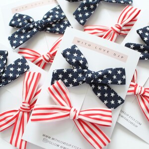 May include: Two sets of hair bows, one set is red and white striped and the other is navy blue with white stars. The bows are displayed on a white card with the text "With Love DJP" and website information.