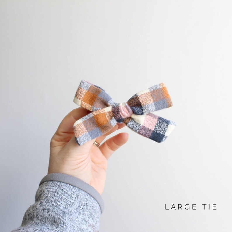 May include: A large plaid bow with a white, pink, blue, and orange color scheme. The bow is tied in a knot and is ready to be worn in hair. The text "LARGE TIE" is visible at the bottom of the image.