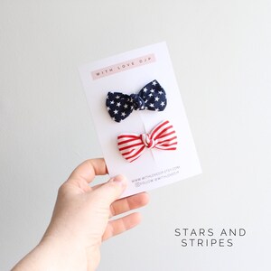 May include: Two hair bows, one with red and white stripes and one with blue and white stars. The bows are on a white card with the text "With Love DJP" and "Stars and Stripes".