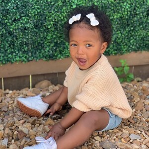 May include: A young child wearing a light pink knitted sweater, denim shorts, and white sneakers. The child has dark brown hair with two white bows in their hair. The child is sitting on a bed of small rocks.