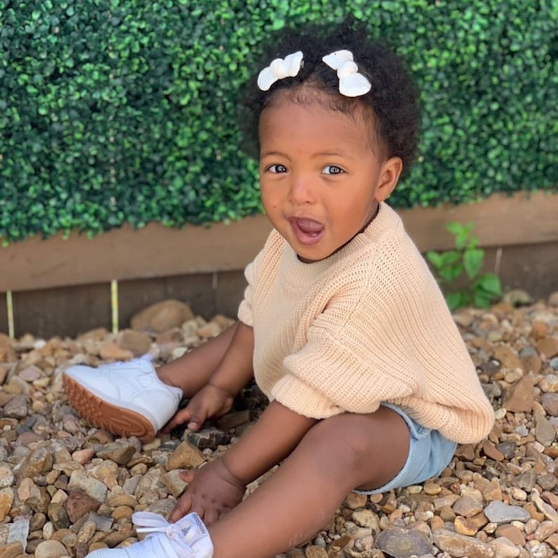 May include: A young child wearing a light pink knitted sweater, denim shorts, and white sneakers. The child has dark brown hair with two white bows in their hair. The child is sitting on a bed of small rocks.