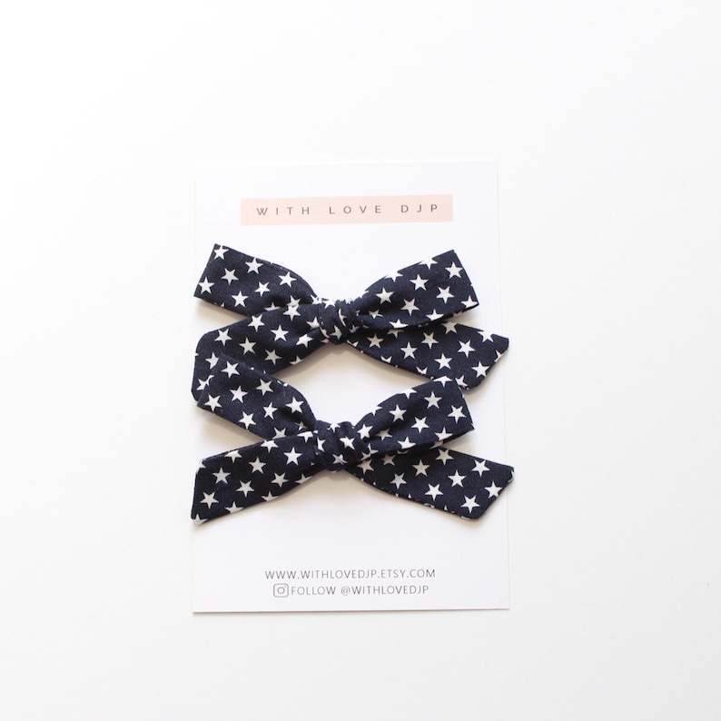 May include: Two navy blue hair bows with white stars. The bows are tied in a knot and are ready to be worn in hair. The bows are on a white background with a business card that says "With Love DJP" and includes a website and social media handle.