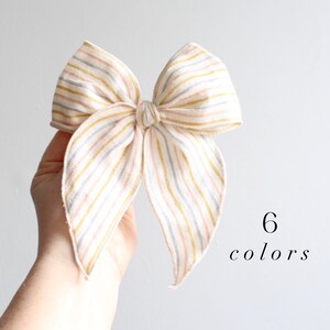 May include: A large, white bow with pink, yellow, and blue stripes. The bow is made of fabric and is tied in a knot. The text "6 colors" is written below the bow.