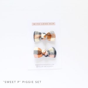 May include: Set of two plaid hair bows in shades of orange, pink, and gray. The bows are tied with a knot in the center and have a "Sweet P" Piggie Set label.