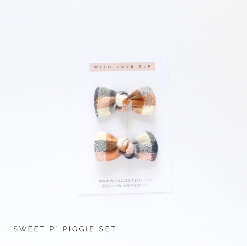 May include: Set of two plaid hair bows in shades of orange, pink, and gray. The bows are tied with a knot in the center and have a "Sweet P" Piggie Set label.
