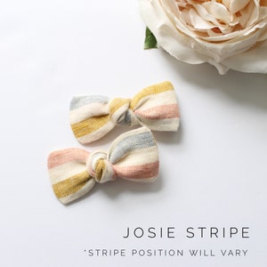 May include: Two striped hair bows in pink, yellow, and gray. The bows are tied in a knot and have a rustic, handmade look. "JOSIE STRIPE" and "STRIPE POSITION WILL VARY" are written below the bows.