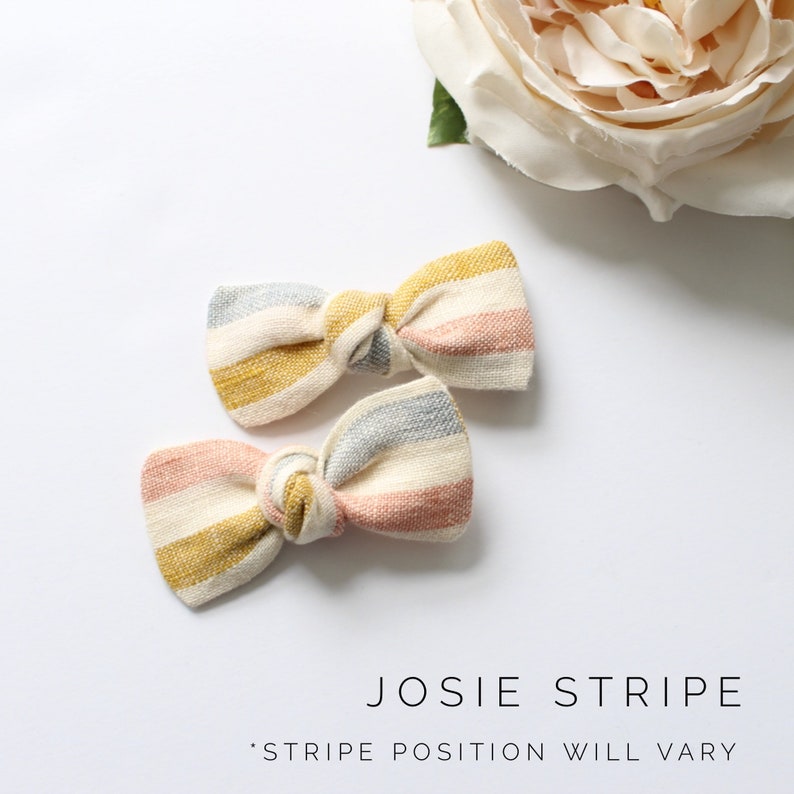 May include: Two striped hair bows in pink, yellow, and gray. The bows are tied in a knot and have a rustic, handmade look. "JOSIE STRIPE" and "STRIPE POSITION WILL VARY" are written below the bows.