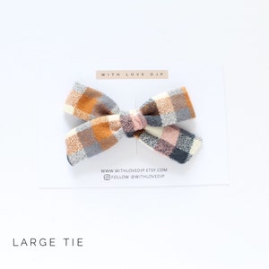 May include: A large plaid bow tie in shades of brown, pink, gray, and white. The bow tie is on a white background with the text "WITH LOVE DJP" and "WWW.WITHLOVEDJP.ETSY.COM" and "FOLLOW @WITHLOVEDJP". The text "LARGE TIE" is at the bottom of the image.