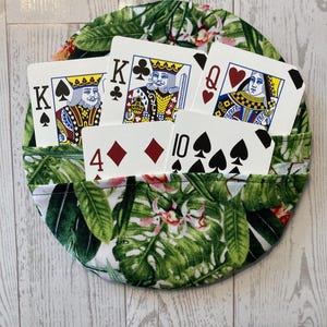 May include: A round playing card holder with a tropical leaf pattern, holding playing cards. The cards visible are the King of Spades, King of Clubs, Queen of Hearts, 4 of Diamonds, and 10 of Spades.