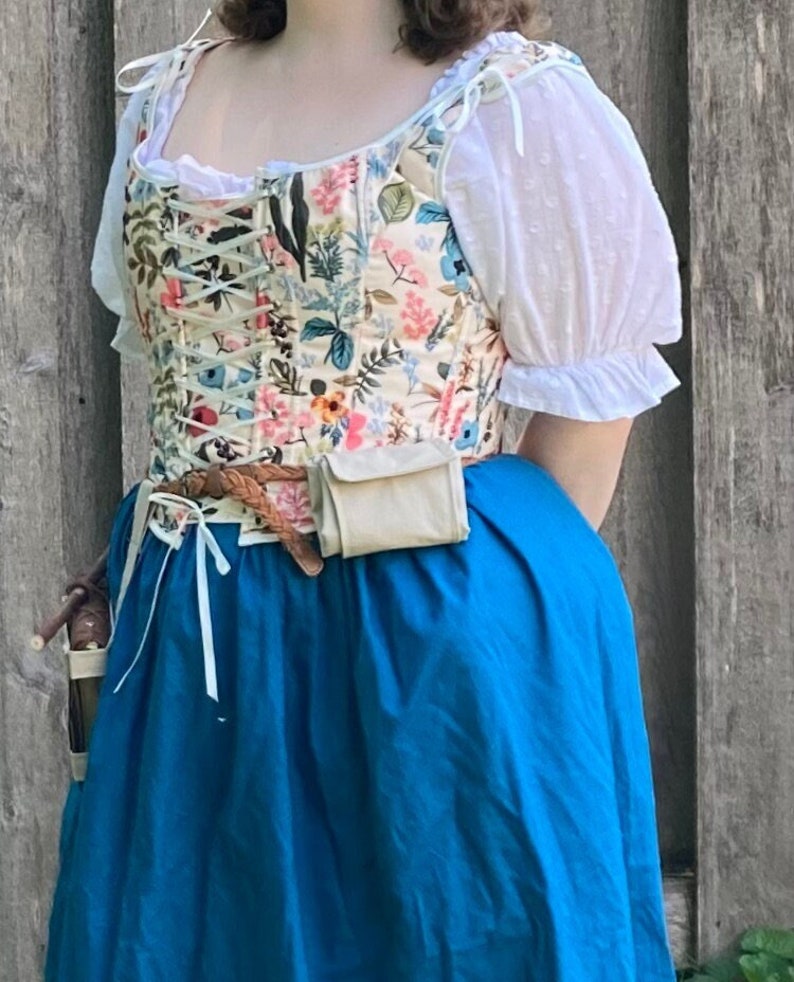 Op de afbeelding: Een bloemenkorset en een blauwe rok. Het korset heeft een cr&egrave;mekleurige achtergrond met roze, blauwe en groene bloemmotieven. De rok is felblauw. Een witte blouse met pofmouwen is zichtbaar. Een bruine riem en een tasje zijn ook aanwezig.