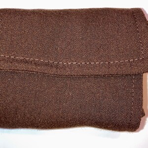 Op de afbeelding: Een kleine, bruine, rechthoekige stoffen etui met een klepsluiting. De etui is gemaakt van een textuurrijk materiaal en heeft zichtbare stiksels langs de randen en de klep. Het algehele ontwerp is eenvoudig en functioneel.