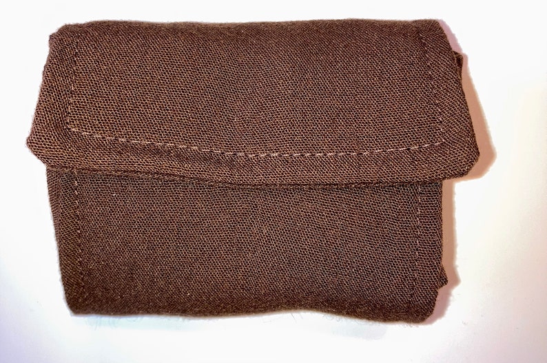 Op de afbeelding: Een kleine, bruine, rechthoekige stoffen etui met een klepsluiting. De etui is gemaakt van een textuurrijk materiaal en heeft zichtbare stiksels langs de randen en de klep. Het algehele ontwerp is eenvoudig en functioneel.