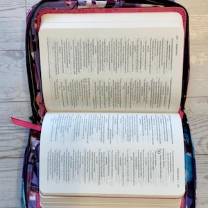 May include: Open Bible in a floral print case with a pink ribbon bookmark. The case is open, revealing the text of the Bible. The case has a purple handle and a zipper closure. The floral pattern includes shades of purple, pink, and blue.