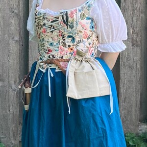 Op de afbeelding: Een bloemenkorset met een cr&egrave;mekleurige achtergrond, geregen aan de voorkant, gecombineerd met een witte blouse en een lange, blauwgroene rok. Een bruine gevlochten riem en een beige zakje met trekkoord maken de outfit compleet.