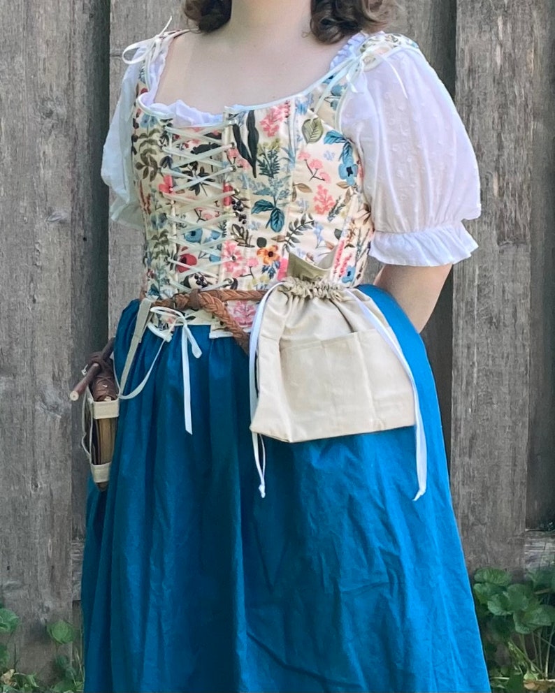 Op de afbeelding: Een bloemenkorset met een cr&egrave;mekleurige achtergrond, geregen aan de voorkant, gecombineerd met een witte blouse en een lange, blauwgroene rok. Een bruine gevlochten riem en een beige zakje met trekkoord maken de outfit compleet.