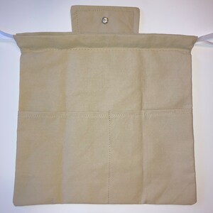 Op de afbeelding: Een beige canvas oprolbare etui met twee rijen zakken en een klepsluiting die is vastgezet met een zilveren drukknop. Witte linten zijn aan de bovenhoeken bevestigd. De etui is ontworpen voor het opbergen van items.