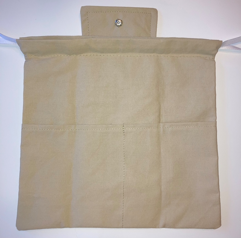 Op de afbeelding: Een beige canvas oprolbare etui met twee rijen zakken en een klepsluiting die is vastgezet met een zilveren drukknop. Witte linten zijn aan de bovenhoeken bevestigd. De etui is ontworpen voor het opbergen van items.