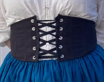 May include: A black corset belt with silver eyelets and white lacing is worn over a white blouse and a blue skirt. The corset cinches the waist, creating a defined silhouette. The outfit suggests a historical or fantasy theme.
