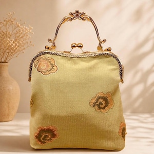 Könnte beinhalten: Eine Handtasche im Vintage-Stil in einem weichen, gedämpften Gelbgrün. Die Tasche hat gestickte Blumenmuster in Braun- und Goldtönen. Sie hat einen dekorativen goldfarbenen Griff und Verschluss, was ihren klassischen Reiz unterstreicht.