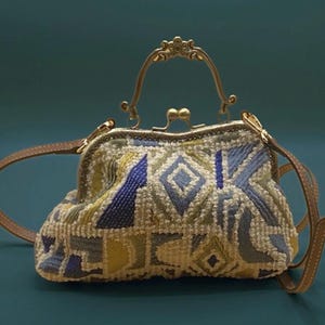 Könnte beinhalten: Eine Handtasche im Vintage-Stil mit einem geometrischen Muster in Blau, Gelb und Weiß. Sie hat einen goldfarbenen Verschluss und Griff sowie einen braunen Lederriemen. Die Tasche hat eine strukturierte Form und eine strukturierte Oberfläche.
