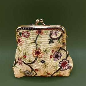 May include: A small, vintage coin purse with a gold-colored metal clasp. The purse is beige with a floral tapestry design in shades of red, purple, and green. The clasp is a kiss-lock style.