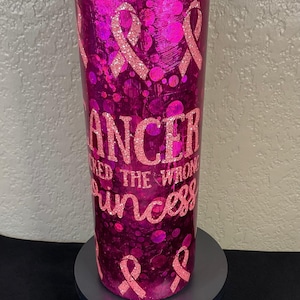 May include: A pink tumbler with a glittery finish, featuring pink ribbon designs and the text "Cancer Picked the Wrong Princess." The tumbler is designed to hold beverages and has a pink lid.