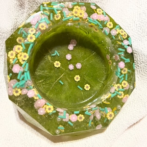 Puede incluir: Un cenicero octogonal con base verde, incrustado con decoraciones florales y de chispas amarillas, rosas y azules. El cenicero tiene un acabado transparente y brillante, sobre un fondo blanco.