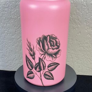 Puede incluir: Un vaso rosa con un diseño de rosa negra. El vaso tiene una tapa negra con una banda iridiscente arcoíris. El vaso cilíndrico está decorado con una rosa negra y hojas. Puede contener aproximadamente 470 ml.