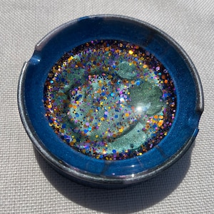 May include: A round, blue ashtray with a metallic silver rim. The interior is filled with a shimmering, multi-colored glitter design, featuring hues of green, gold, purple, and blue. The ashtray is likely made of resin or a similar material.