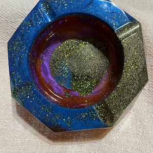 Puede incluir: Un cenicero octogonal con un diseño de resina brillante azul, morado y dorado. El cenicero tiene un interior marrón oscuro y un exterior gris oscuro. El cenicero es un artículo decorativo.