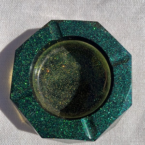 May include: An octagonal ashtray with a glittery green exterior and a circular interior. The centre of the ashtray has a gold and green glitter design. The ashtray is made of a clear material.