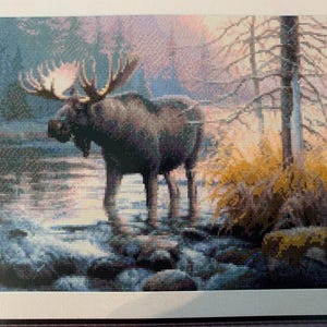May include: Framed artwork featuring a majestic moose standing in a stream. The moose has large antlers and is surrounded by trees and foliage. The artwork uses a pixelated style with a color palette of blues, browns, and yellows.