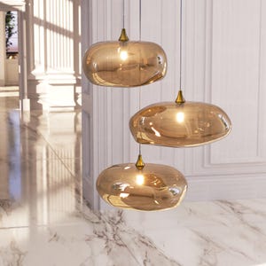 May include: A cluster of three pendant lights featuring irregular, amber-tinted glass shades with a reflective, metallic finish. Each shade is suspended by a thin wire from a small, conical brass-coloured fixture, creating a warm, glowing effect.
