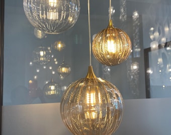 Op de afbeelding: Drie glazen hanglampen met geribbelde texturen. De bovenste lamp is van helder glas, de middelste lamp is van amberkleurig glas en de onderste lamp is van donkerder amberkleurig glas.