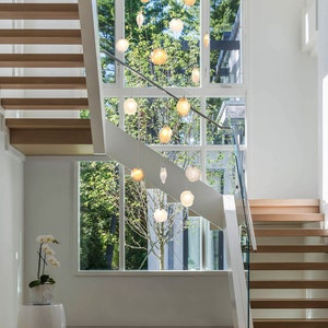 May include: A modern staircase with wooden steps and a glass railing. A large window with a view of trees and a chandelier with multiple light bulbs hangs from the ceiling.