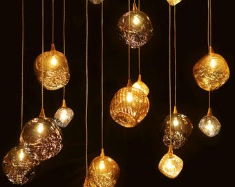 May include: A collection of 13 gold and brown glass pendant lights hanging from the ceiling. The lights are all different shapes and sizes, and they are all lit up.