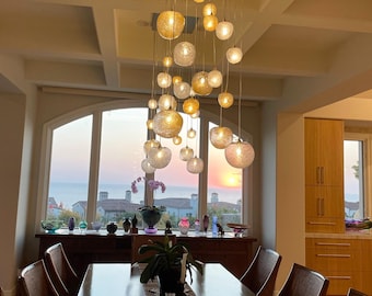 Op de afbeelding: Een moderne eetkamer met een groot raam met uitzicht op de oceaan. Een kroonluchter met meerdere glazen bollen hangt boven een houten eettafel met stoelen. De kroonluchter bestaat uit een mix van heldere en amberkleurige glazen bollen.