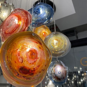 Puede incluir: Una lámpara de araña con múltiples orbes de vidrio en tonos de azul, naranja y rojo. Los orbes tienen un patrón de remolino y están suspendidos del techo por cables finos.