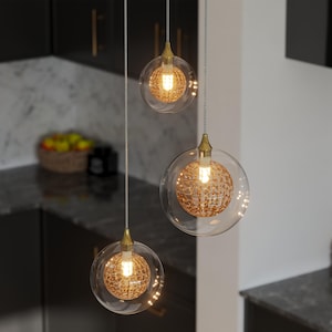 May include: Three pendant lights with clear glass globes and gold-coloured accents. Each globe houses a textured, spherical light fixture that emits a warm glow. The lights hang from thin, silver cords, creating a modern and elegant aesthetic.