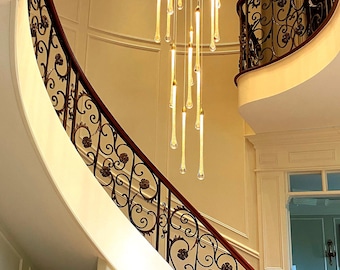 May include: A modern, gold-toned chandelier with long, thin, teardrop-shaped glass pendants hangs from the ceiling of a grand staircase with ornate wrought iron railings.